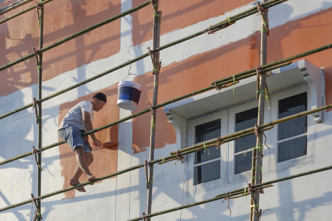 Asiatischer Handwerker streicht eine Hausfassade bei sonnigem, trockenem Wetter