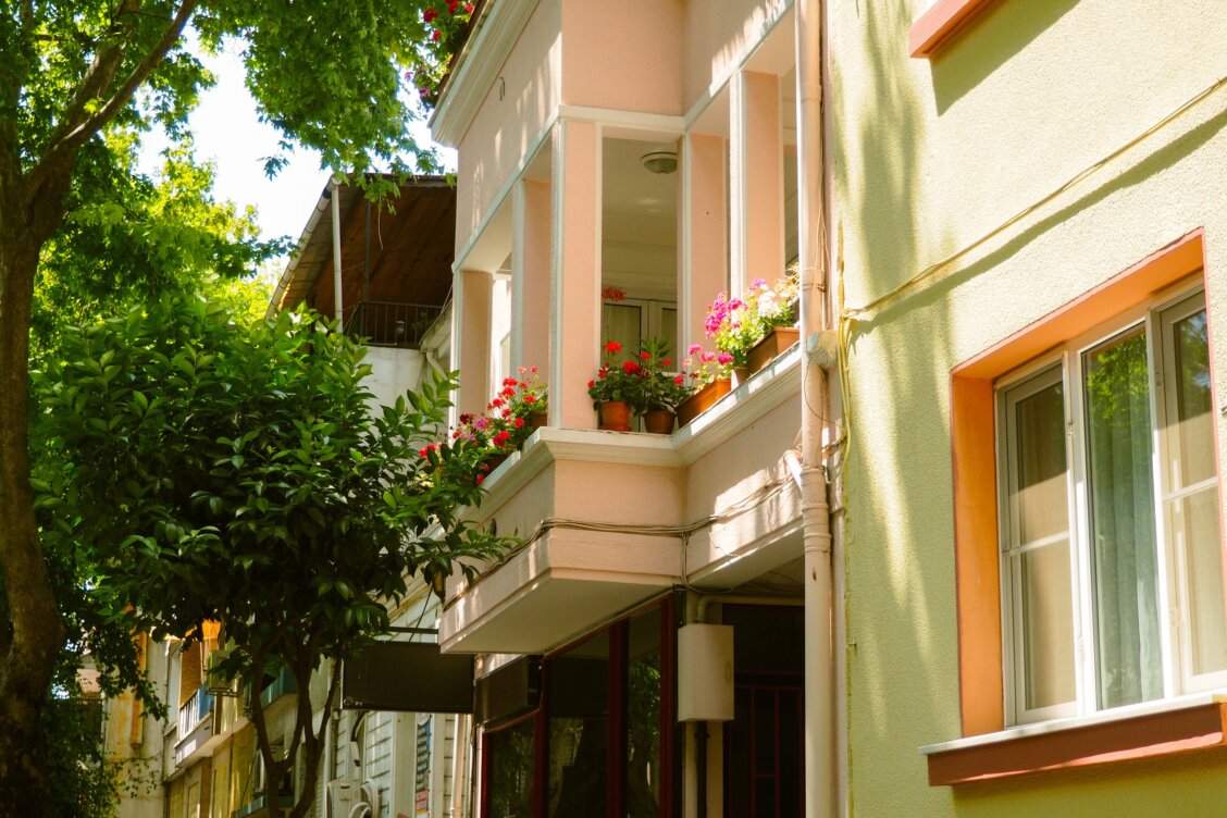 Bild von Edanur Sonkaya auf Pexels: Balkon mit Blumenkästen und Töpfen an einem Wohnhaus, teils in Sonne, teils im Schatten von Bäumen