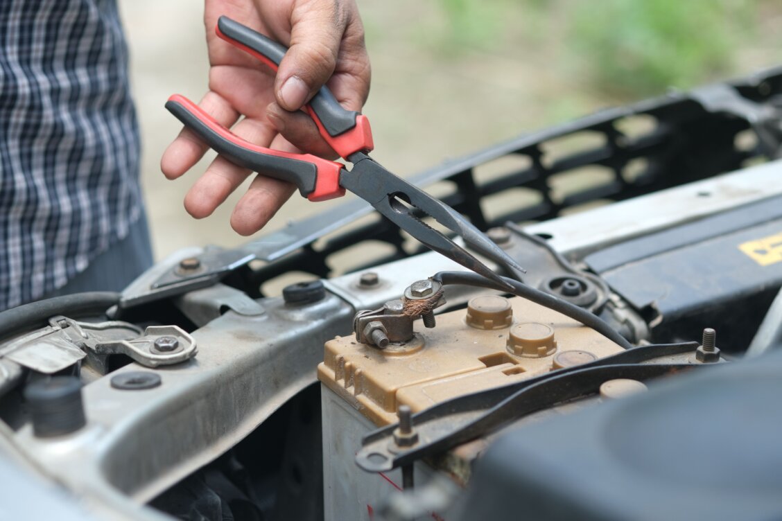 Bild von Towfiqu barbhuiya auf Pexels: Eine Hand hält eine Zange über den Polen einer Autobatterie im geöffneten Motorraum