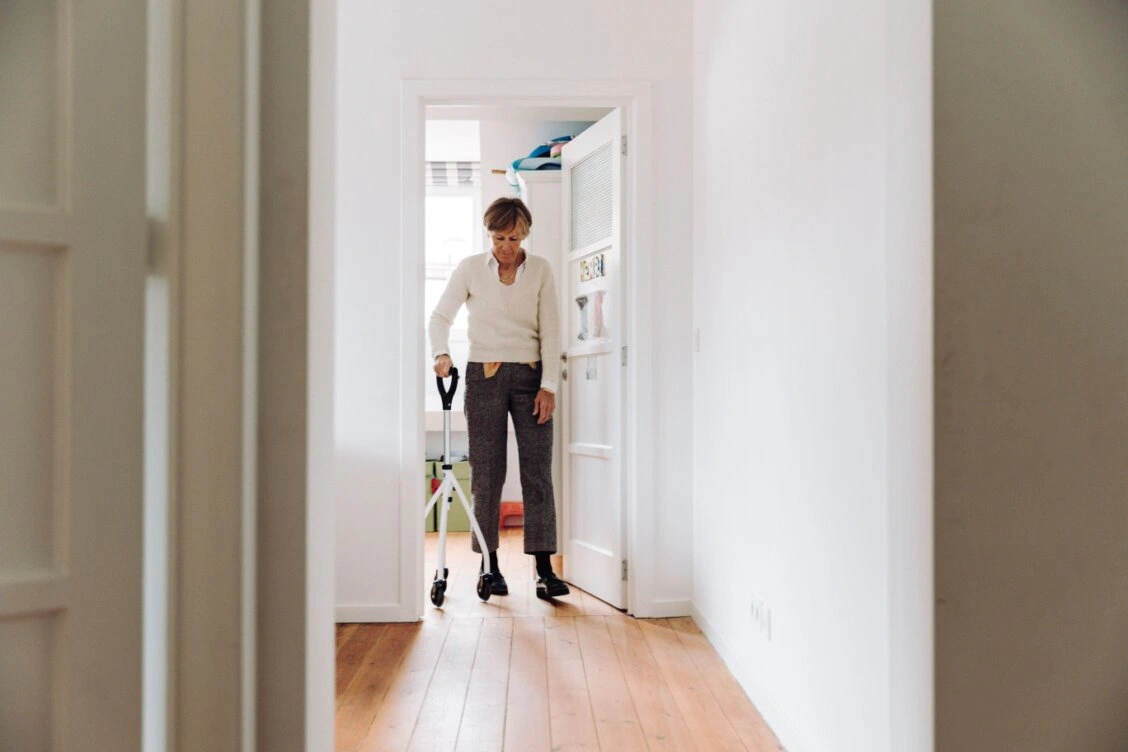 Bild von Wheeleo Walker auf Pexels: Eine ältere Frau geht in einem hellen Flur zu Hause mit einem Gehhilfswagen.
